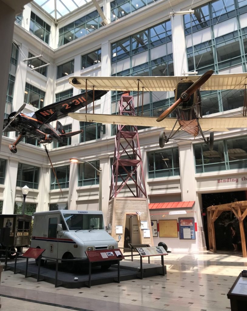 photo of exhibit hall at the National Postal Museum in Washington DC, showing 2 planes used for early airmail, and a modern delivery truck.