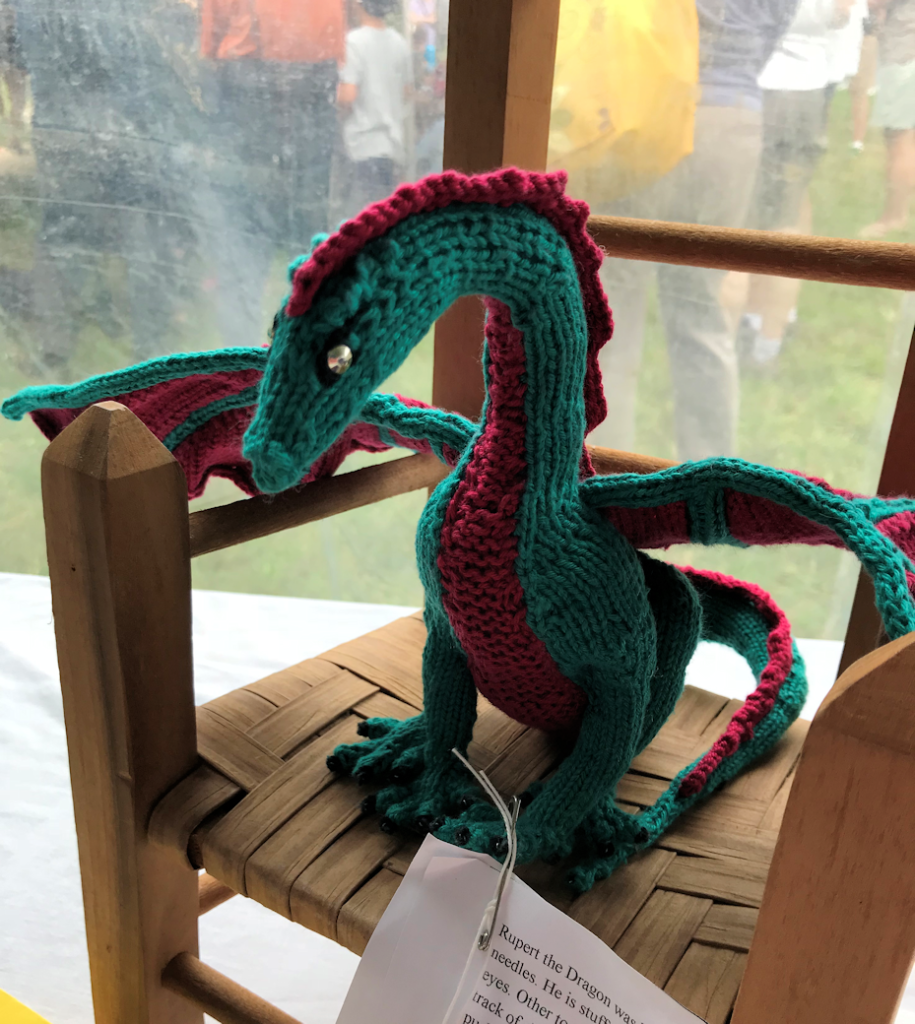 knitted dragon in burgundy and dark green, sitting on miniature woven-seat wooden chair