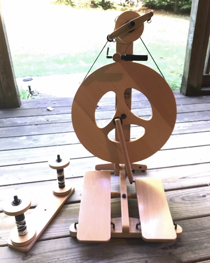 A Louet Victoria S95 spinning wheel and lazy kate with two empty bobbons. They are sitting on a  weathered front porch. The Victoria is a double-treadle castle wheel (an upright wheel, with the flyer above the wheel); it has a modern aesthetic. The circular wheel itself looks like a peace sign, with three oval cutouts.